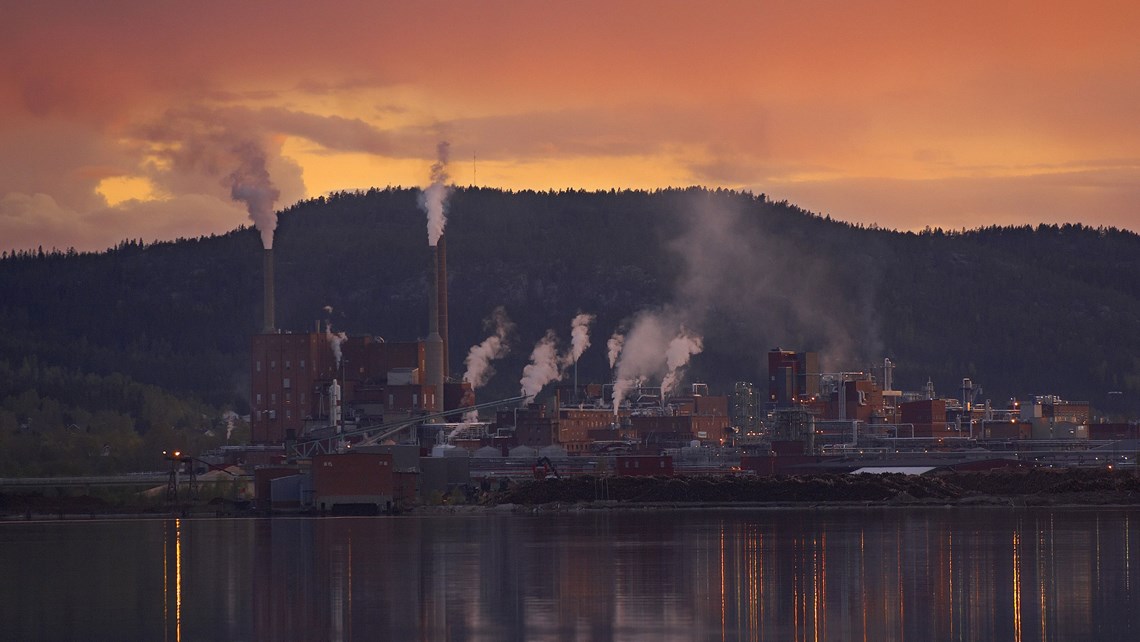 Sonnenuntergang über dem Domsjö-Werk vor einer Waldkulisse und einem Fluss im Vordergrund.